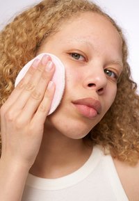 Un disque de coton rond et rose clair est appliqué sur un visage avec des cheveux blonds bouclés. La peau présente un teint naturel avec quelques imperfections légères.