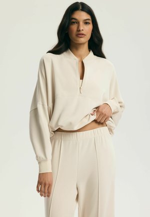 Kobieta w kremowej, zapinanej na zamek bluzie z długim rękawem i dopasowanych spodniach, lekko podciągająca bluzę, odsłaniając talię.