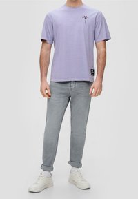 Camiseta de manga corta lila con un gráfico de flor en el lado izquierdo del pecho, combinada con jeans gris claro y zapatillas blancas.