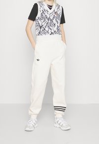 Bílé oversized tepláky se černými pruhy, v kombinaci s texturovanou bílou vestou s černým vzorem přes černé tričko. Bílé tenisky.