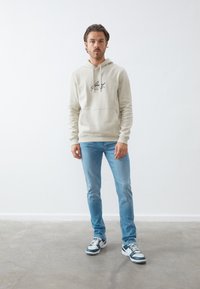Lys beige hættetrøje med et sort logo, parret med lyseblå jeans og flerfarvede sneakers, stående på en neutral baggrund.