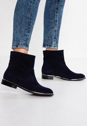 Person iført tætsiddende blå jeans og mørkeblå ruskindsstøvler med lave hæle, stående på hvidt gulv.