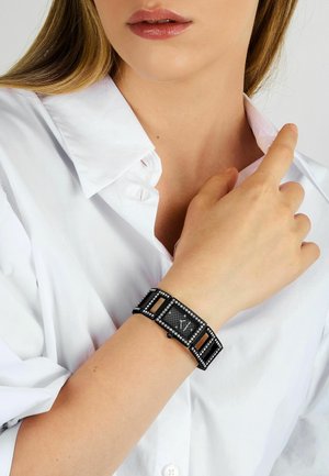Black watch with rectangular face, diamond-like accents, and a textured band. Worn on a wrist, paired with a white collared shirt.