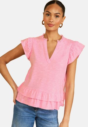 Frau trägt eine pinke Kurzarmbluse mit Spitzen-Details und Rüschen, kombiniert mit blauen Jeans, posiert mit einer Hand auf der Hüfte.