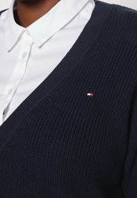 Marinblå rippad stickad cardigan med djup V-hals, strukturerad textil och liten röd, vit och blå logotyp på bröstet. Vit skjorta med krage under.
