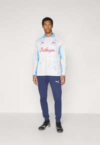 Maglietta a maniche lunghe di colore azzurro chiaro con stampa floreale e testo rosso "KidSuper", abbinata a pantaloni della tuta blu navy e scarpe da ginnastica nere.