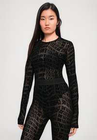 Juodas tinklelio bodysuit su ilgomisRankovėmis, su viso paviršiaus logotipo atspaudu. Lygus paviršius, prigludusi forma ir firminis liemens juostos dizainas.