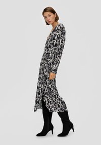 Leopardenmuster-Kleid in grauen Tönen, mit langen Ärmeln und tiefem V-Ausschnitt. Der Stoff ist glatt mit einem fließenden Design und Seitenschlitz. Getragen mit schwarzen Stiefeletten mit Absatz.