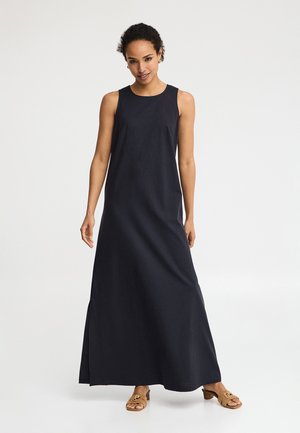 Kobieta stojąca w czarnej, bezrękawnikowej sukni maxi i brązowych, otwartych sandałach na niskim obcasie na jednolitym białym tle.