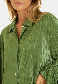 Camisa verde texturizada con un patrón en forma de ola, tejido brillante, cierre de botones y cuello, con un corte relajado y mangas largas.