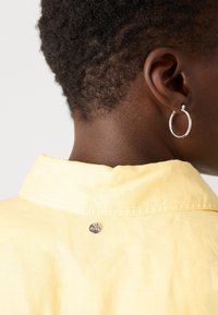 Gros plan sur le cou et l’oreille d’une personne aux cheveux courts et bouclés, portant une boucle d’oreille argentée en forme d’anneau et une chemise col jaune clair.
