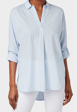 Chemise rayée bleu clair et blanc à manches longues avec col, manches retroussées et un pli devant, portée avec un pantalon blanc.