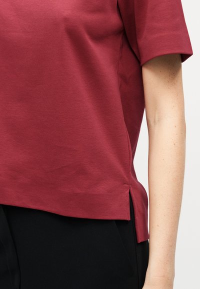 Personne portant une chemise rouge à manches courtes avec une fente latérale et un pantalon noir, montrant les détails du bras droit et du torse.