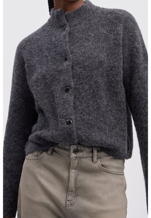 Person iført en fuzzy mørkegrå knapstukket cardigan, der er stoppet ned i lysebeige højtaljede jeans med synlige bæltesløjfer og lommer.