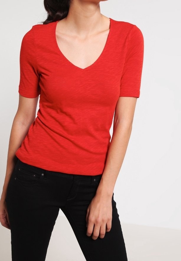 Femme portant un t-shirt rouge ajusté à col en V et manches courtes, et un pantalon noir, debout devant un fond clair uni.