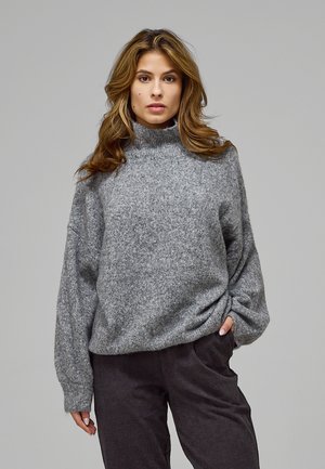 Femme aux cheveux châtain clair portant un pull à col roulé gris et un pantalon foncé, debout devant un fond gris uni.