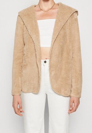 Beige, blød åben-front jakke båret over en hvid tube top og højtaljede hvide bukser, med en synlig guldkæde.