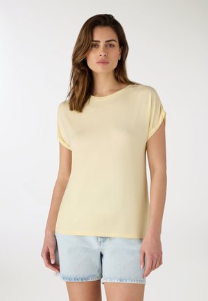 Donna con capelli castani alla lunghezza delle spalle che indossa una maglietta a maniche corte di colore giallo pallido e pantaloncini di denim blu chiaro, in piedi su uno sfondo semplice.