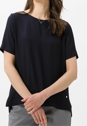 Kvinde iført en ensfarvet sort top med korte ærmer, mønstrede bukser, et delikat halskæde og en tynd ring med hænder foldet foran.