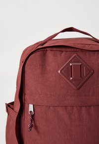 Bordeaux ryggsekk med tekstiloverflate, som har et diamantformet logo, glidelås lukking, og en sidelomme med snor til glidelåsen.