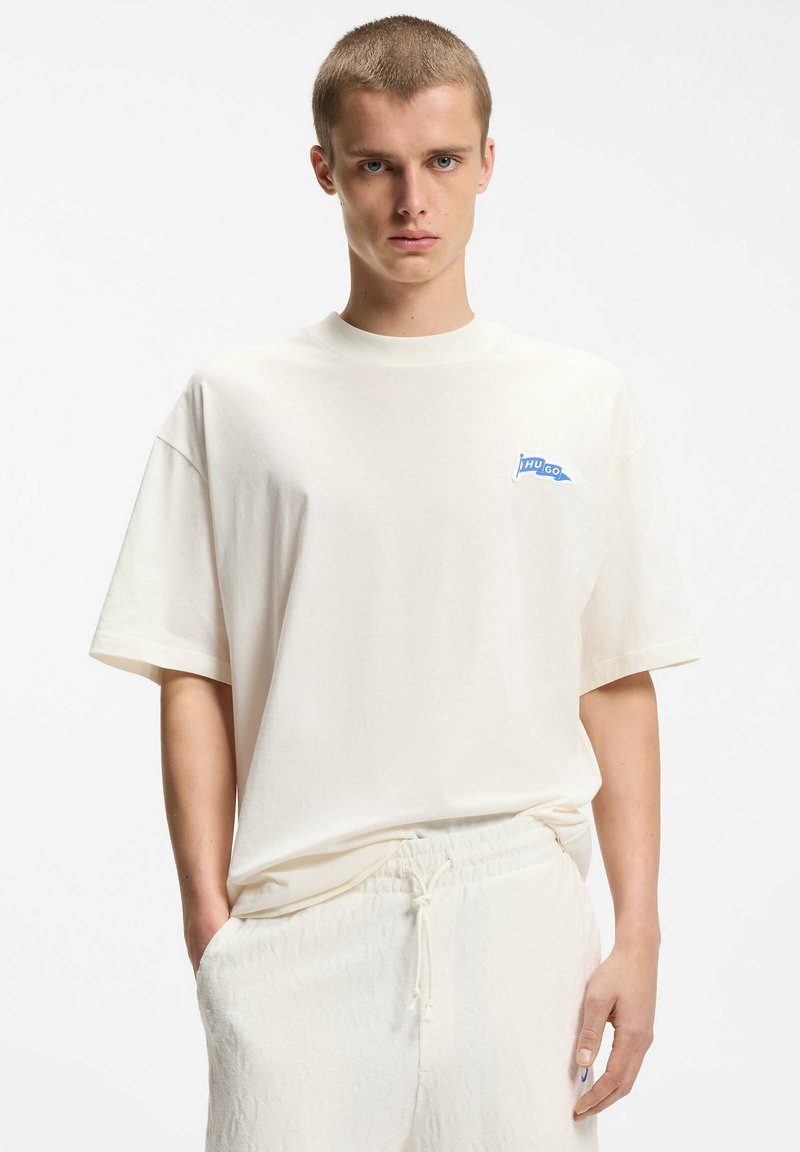 Jeune homme aux cheveux courts portant un t-shirt blanc ample avec un petit logo bleu "HUGO" et un pantalon blanc à cordon de serrage, sur un fond uni.