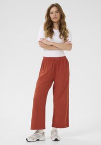 Pantalons larges en tissu orange rouille avec une taille élastique, assortis à un haut blanc ajusté et des baskets de couleur claire.
