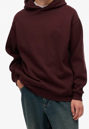 Person trägt einen dunkelburgunderfarbenen Kapuzenpullover mit kleinem Logo auf der Brust und blaue Jeans, eine Hand in der Tasche, vor neutralem Hintergrund.