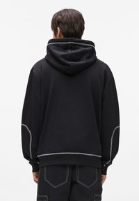 Melna kokvilnas kapuce ar lielu kapuci, kontrastējošām pelēkām šuvēm un brīvu piegriezumu. Iezīmēta ar noapaļotu apakšmalu un brīvām piedurknēm.