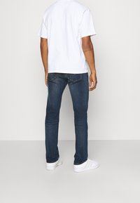 Hombre con vaqueros azul oscuro, zapatillas deportivas blancas y una camiseta blanca sencilla de espaldas frente a un fondo claro.