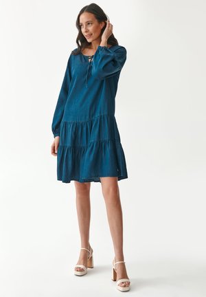 Denim-tiered dress in dark blue with long sleeves and a lace-up neckline. Features a loose fit and flared bottom tiers, paired with strappy heels.