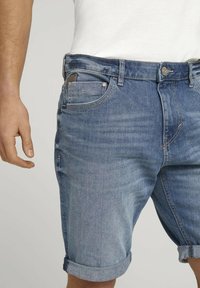 Pantalones cortos de mezclilla azul con dobladillo enrollado, que cuentan con un bolsillo frontal, lazos para el cinturón y un sutil patrón de lavado. El material parece suave y flexible.