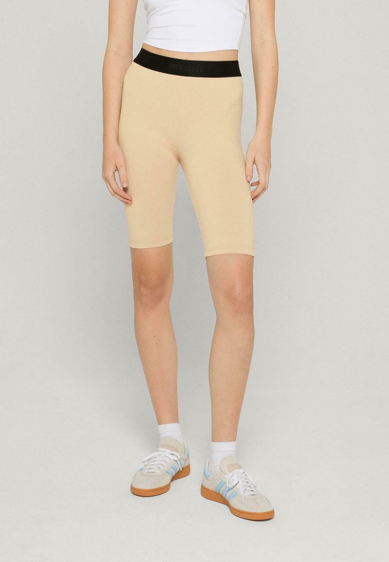 Béžové cyklistické šortky vyrobené z elastického materiálu, s čiernym elastickým pásom so logom. Hladký povrch, dizajn do dĺžky po kolená.