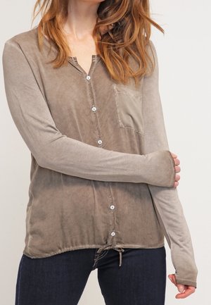 Kvinde iført en taupefarvet skjorte med knapper, lange ærmer, frontlomme og snøre i kanten, kombineret med mørkeblå jeans.