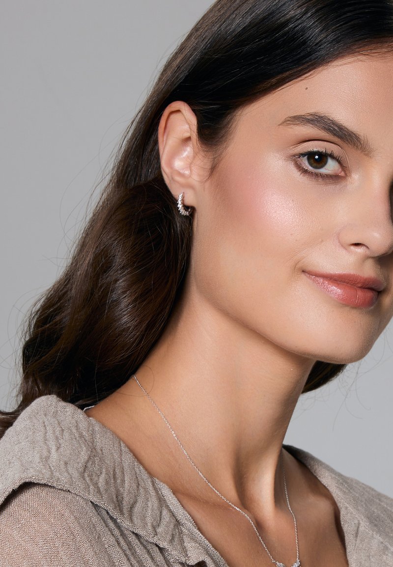 Srebrna kolczyk w kształcie obręczy z kryształowymi akcentami, o zakrzywionym kształcie. Modelka nosi teksturowany, jasny top oraz delikatny naszyjnik.