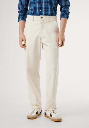 Mężczyzna w prostych, kremowych spodniach, niebieskiej koszuli w kratę oraz białych trampkach z czarnymi paskami i gumową podeszwą, stoi na białym tle.