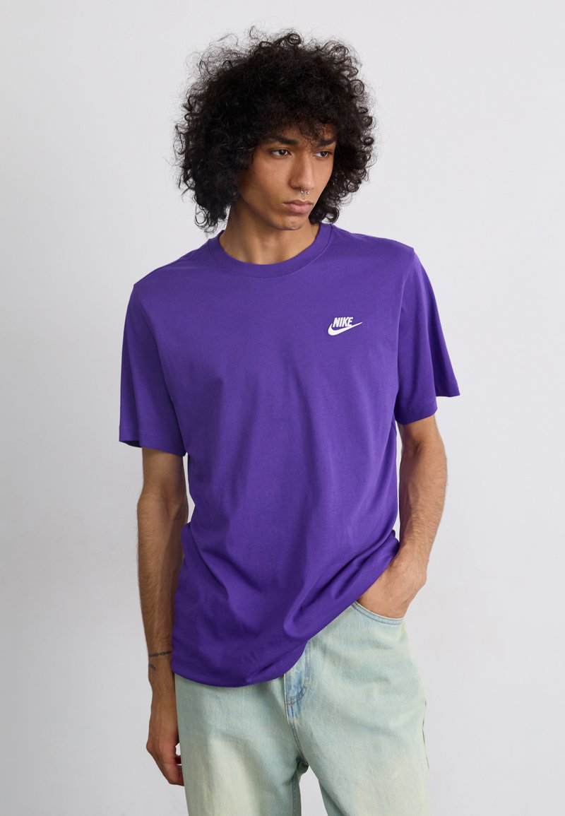 Lilla bomulds t-shirt med korte ærmer, rund hals, og hvid Nike-logo på venstre bryst. Bæres med lyseblå denim jeans.