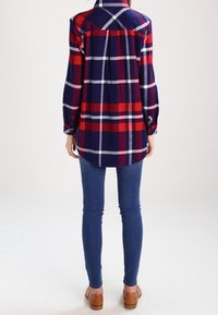 Rutig skjorta i röd, marinblå och vit, med avslappnad passform, långa ärmar och spetsig krage. Matchas med blå jeans och bruna loafers.