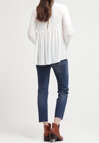 Biała bluzka z długim rękawem i marszczoną talią, z odkrytymi plecami, zestawiona z niebieskimi wąskimi dżinsami i brązowymi botkami za kostkę z teksturowanym wykończeniem.