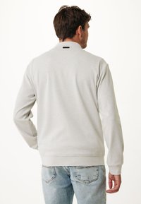 Světle šedý svetr s dlouhými rukávy, ribbenými manžetami a lemem, s malou černou nášivkou pod límcem. Nosič má na sobě modré džíny.