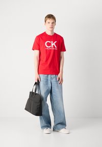 Raudona medvilninė marškinėliai su baltais užrašais "CK CALVIN KLEIN", derinami su šviesiai mėlynomis plačiomis kelnėmis ir juoda rankine, laikoma už rankenos.