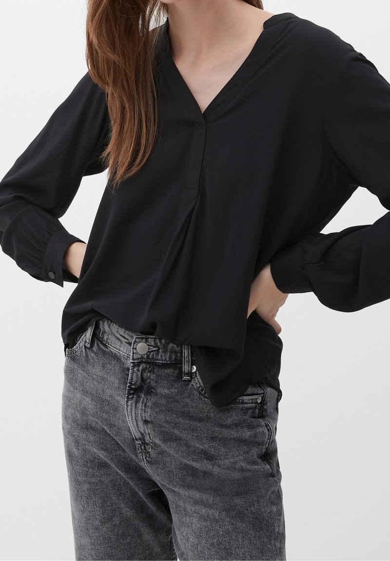 Blouse noire à col en V avec manches longues et poignets à revers, associée à un jean en denim gris avec texture délavée et détail bouton.