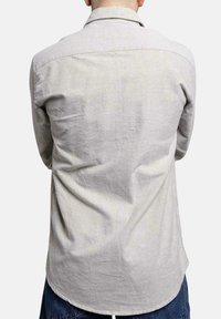 Camisa gris clara hecha de una tela suave, con un cuello clásico, mangas largas y un dobladillo curvado. Detalles de costura visibles en los hombros.