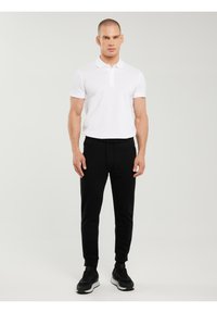 Mees seisab seljas valge lühikeste varrukatega polo särk, musta nööriga dressipüksid ja mustad tossud lihtsa tausta ees.