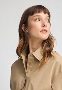 Beige, knöpfbares Hemd mit spitzem Kragen und glatter Textur. Lockerer Schnitt mit sichtbaren Knöpfen und einem entspannten, lässigen Design.
