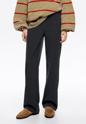 Personne portant un pantalon gris foncé à jambes larges, des mocassins marron clair avec doublure en fourrure, et un pull beige à rayures rouges.