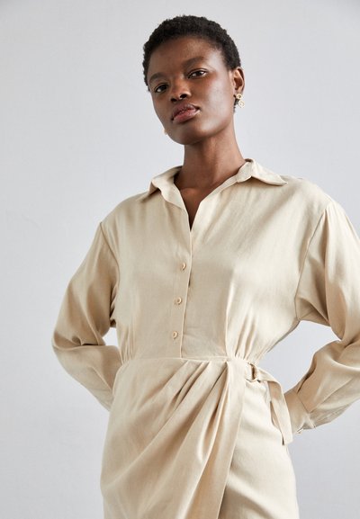 Vestido beige de botones, con mangas largas, que presenta un cuello y un detalle de lazo en el lado. Está hecho de una tela suave con una textura drapeada.