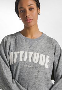 Sudadera gris con textura suave, con la palabra "ATTITUDE" impresa en letras blancas desgastadas y cuello redondo con mangas raglán.