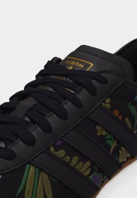 Czarny sneaker Adidas z czarnymi sznurowadłami, złotym logo na języku i kwiatowym haftem w kolorach zielonym, żółtym, fioletowym i pomarańczowym po bokach.