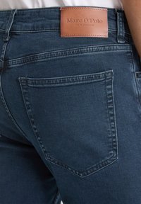 Gros plan sur un jean en denim bleu foncé montrant la poche arrière et l'étiquette de marque en cuir marron "Marc O’Polo" cousue sur la ceinture.