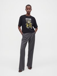 Γυναίκα που φοράει μαύρο t-shirt Moschino με σχέδιο αρκουδάκι, γκρι φαρδιά παντελόνια και μαύρα πέδιλα με ανοιχτά δάχτυλα, στέκεται με τα χέρια στις τσέπες.